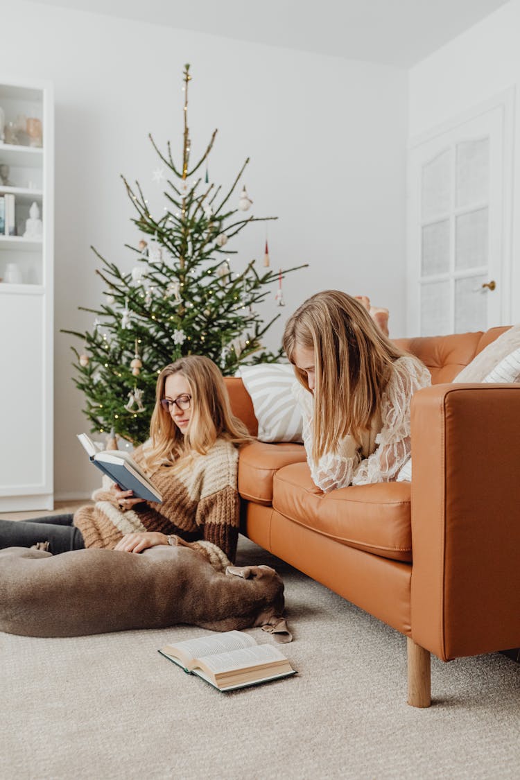 Woman And A Girl Reading Books In A Living Room With Christmas Tree And Dog Lying On Floor