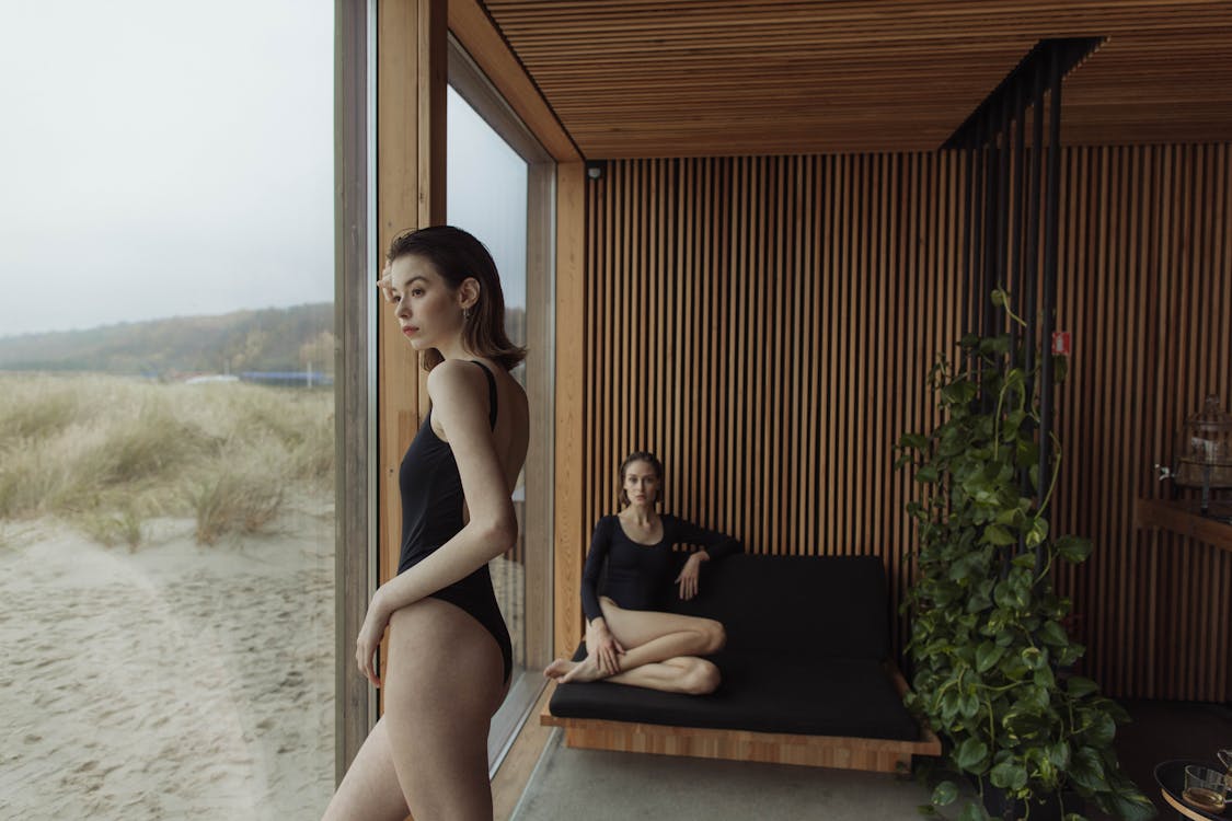  A Woman Wearing Swimwear Standing Beside the Grass Wall While Another Woman Sitting on the Couch 