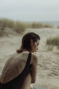 A woman in a black swimsuit sits pensively on a sandy beach with grass.