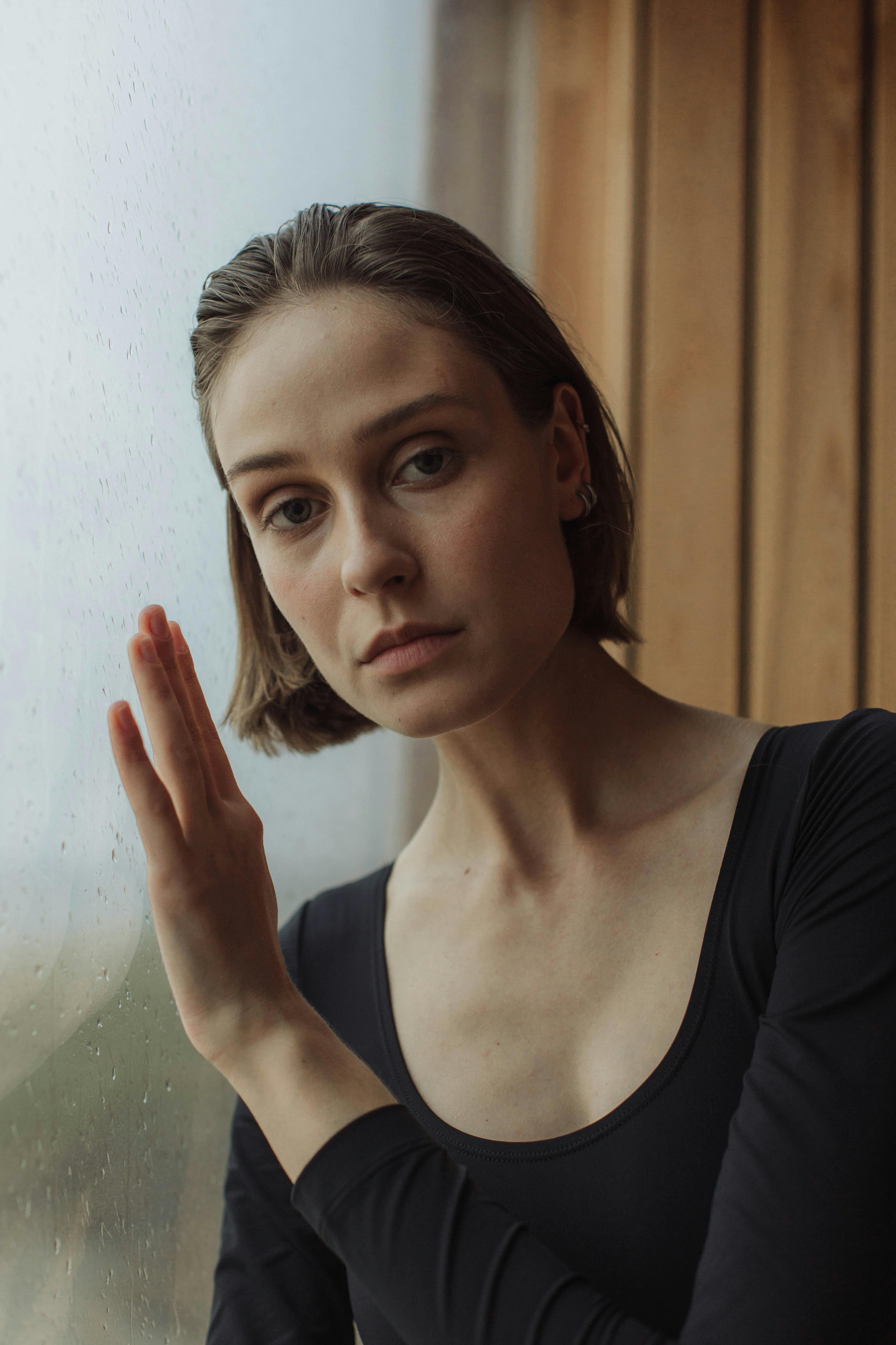 A woman in a black outfit leans on a glass wall, captured in a moody portrait with soft lighting.