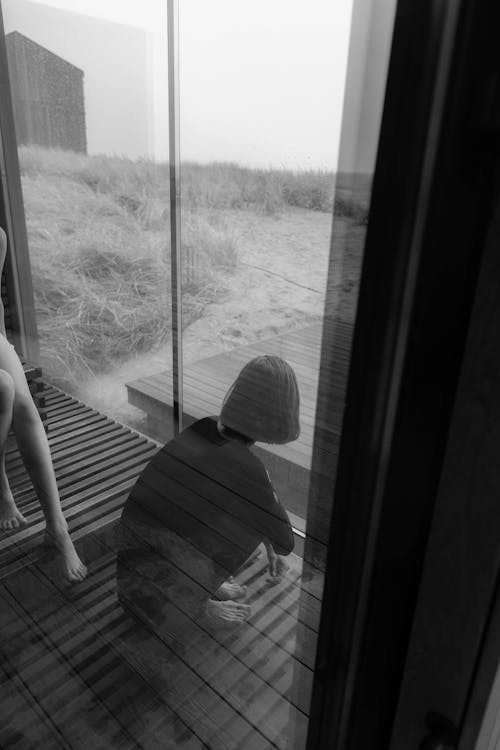 Grayscale Photo of Woman Sitting on the Floor Near the Glass Wall
