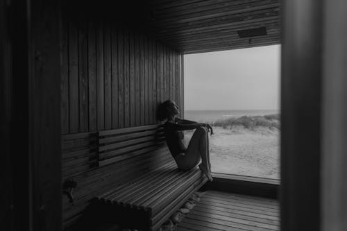 Grayscale Photo of Woman Sitting on a Wooden Bench Near the Glass Wall 