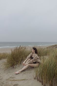 一名妇女坐在沙滩的草丛中，在多云的天空下凝视着宁静的海平线