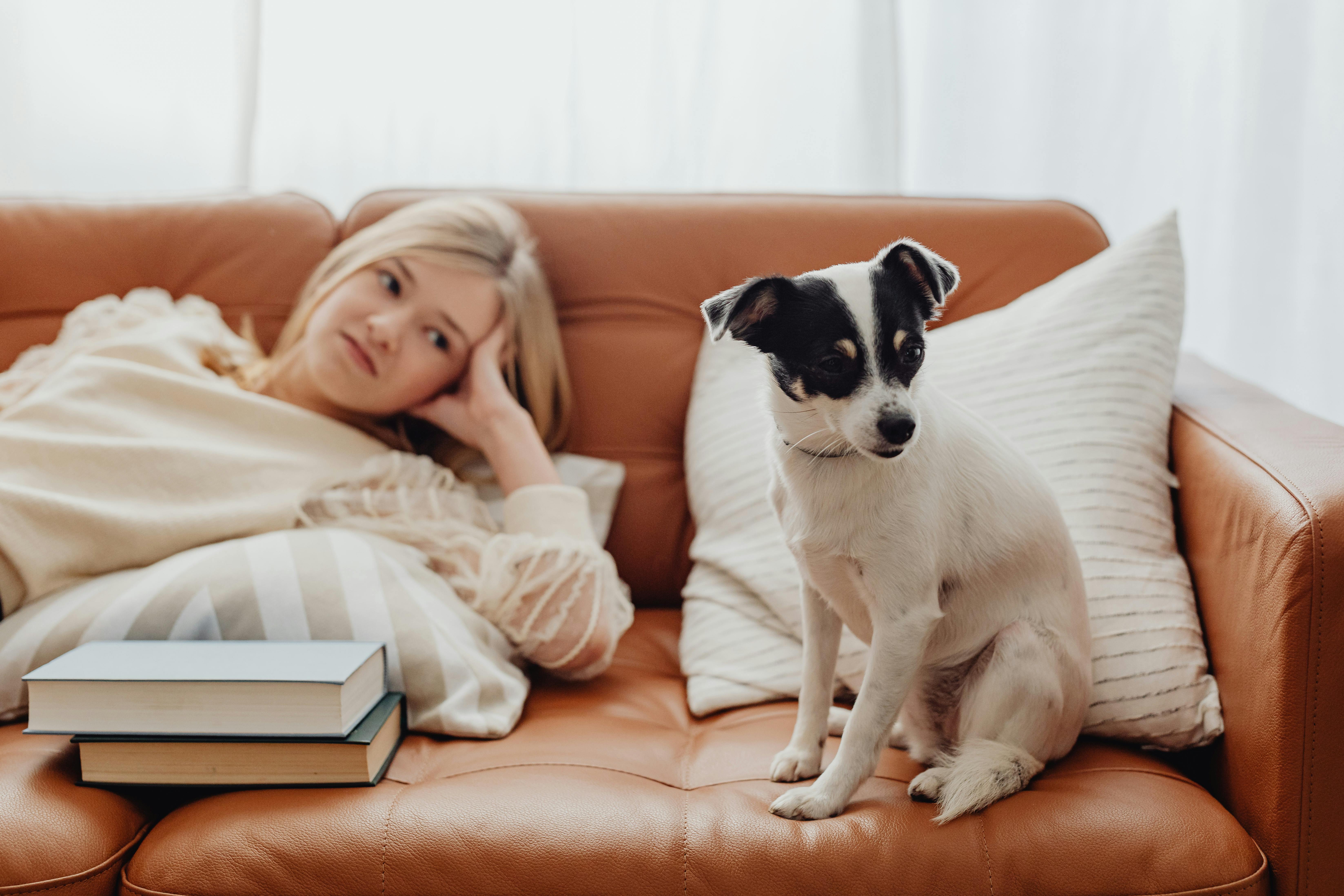 A young woman and her dog relax on a leather sofa surrounded by books.