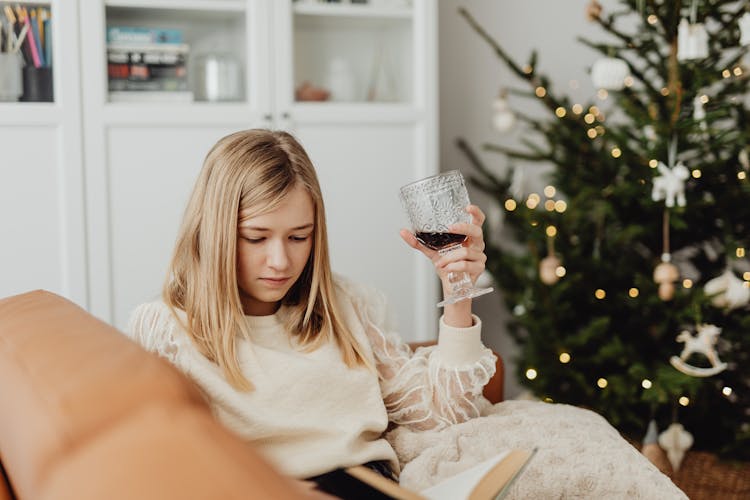 Woman Sitting Reading A Book And Drinking Wine With A Christmas Tree In The Background 