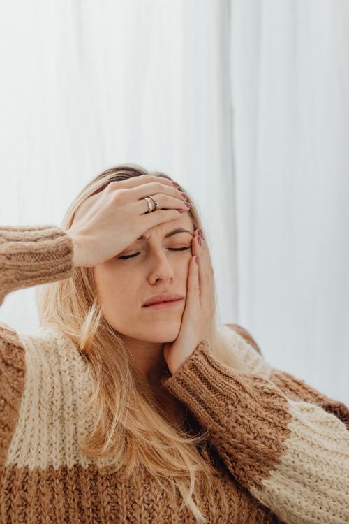 Free Woman with closed eyes holding head, wearing a knitted sweater, possibly experiencing discomfort. Stock Photo