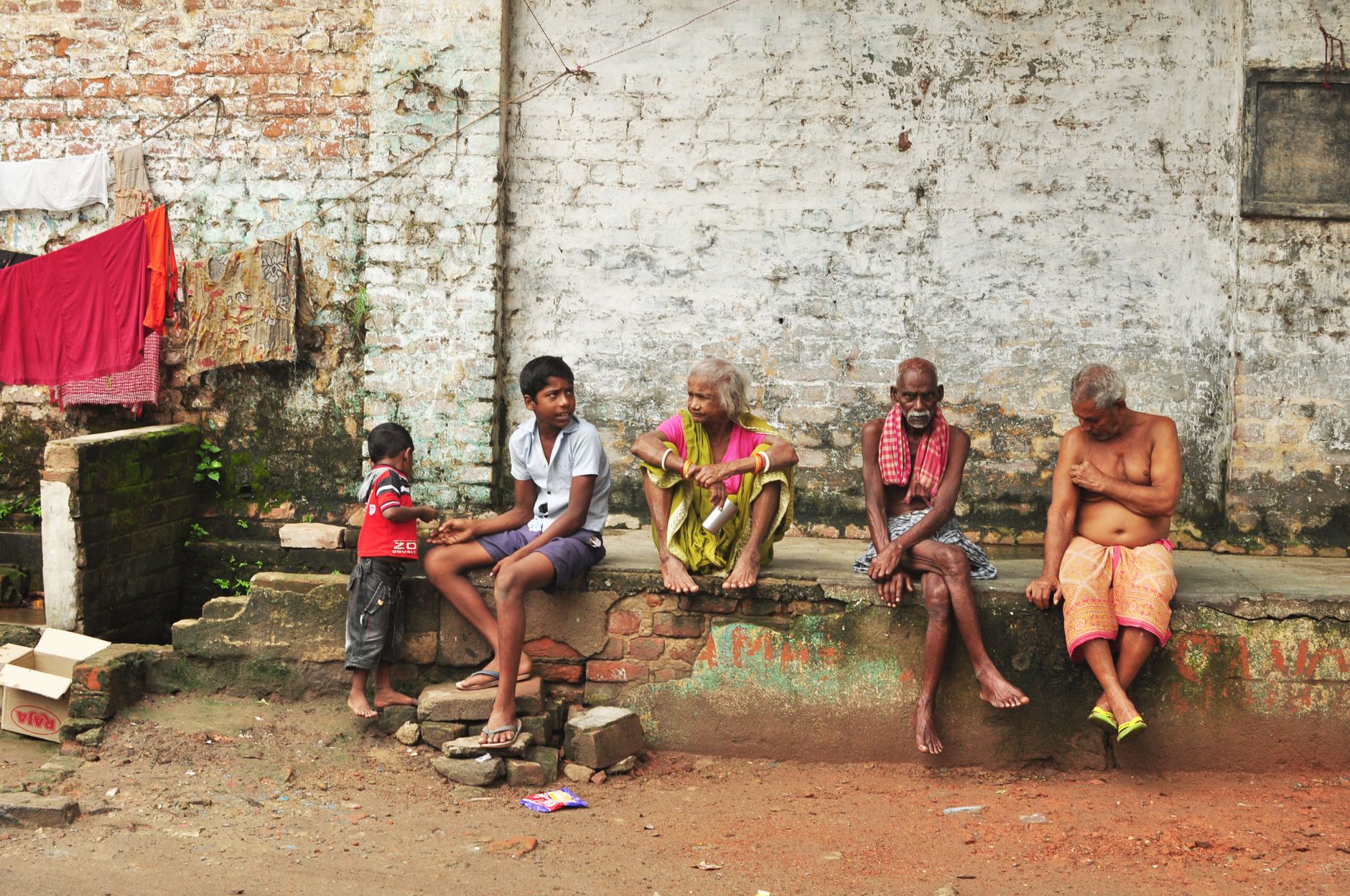 A candid street photo featuring a group of people sitting against a brick wall in Baranagar, India.