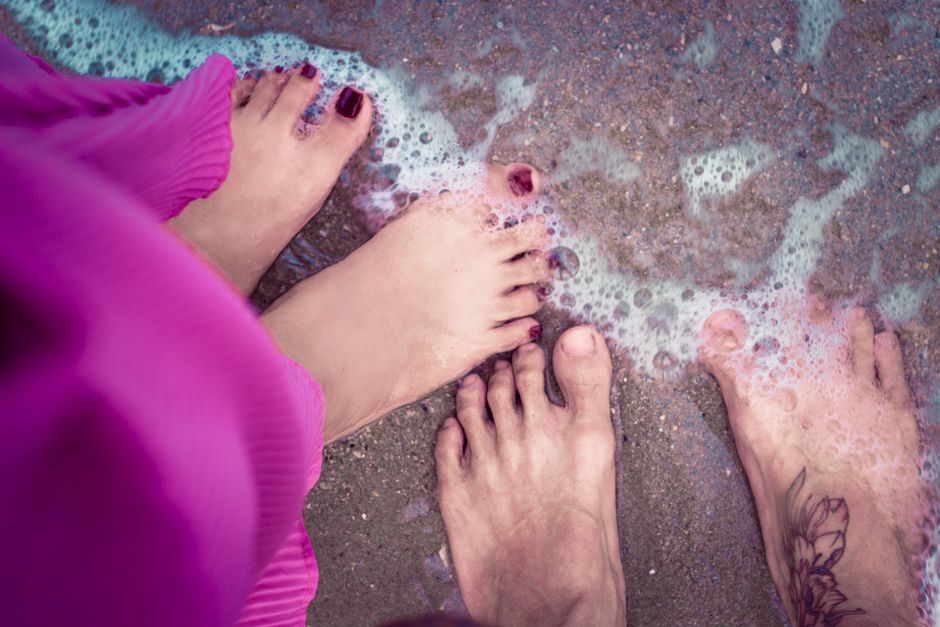 Three pairs of feet in ocean waves with sandy beach and purple attire.