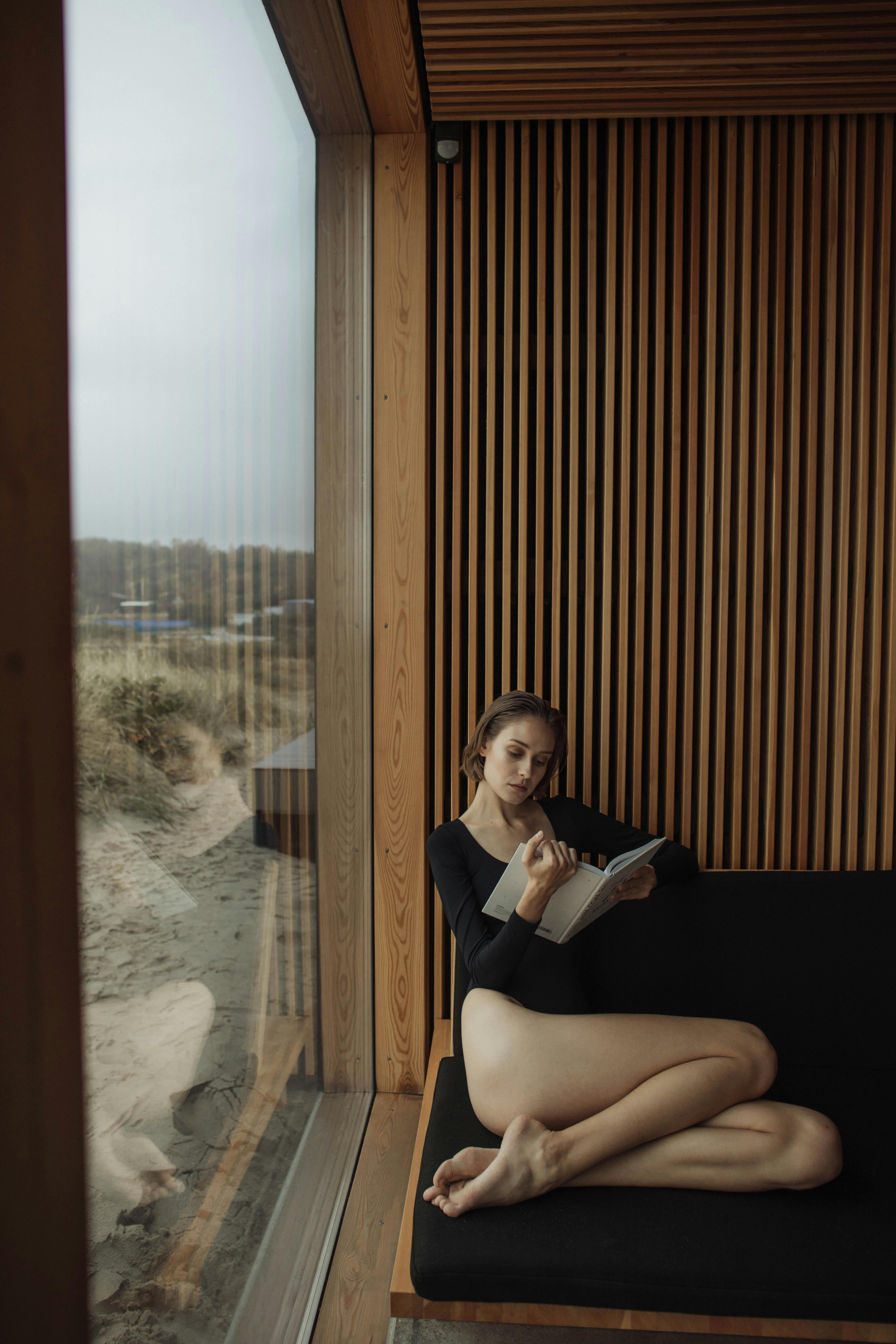 Free Woman reading a book on a sofa by a glass wall with a beach view, creating a serene indoor atmosphere. Stock Photo