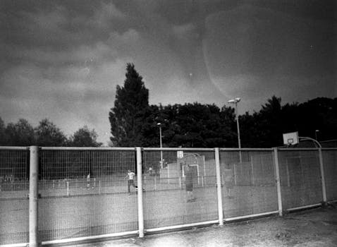Black and white image of an outdoor basketball court surrounded by a fence on a cloudy day.