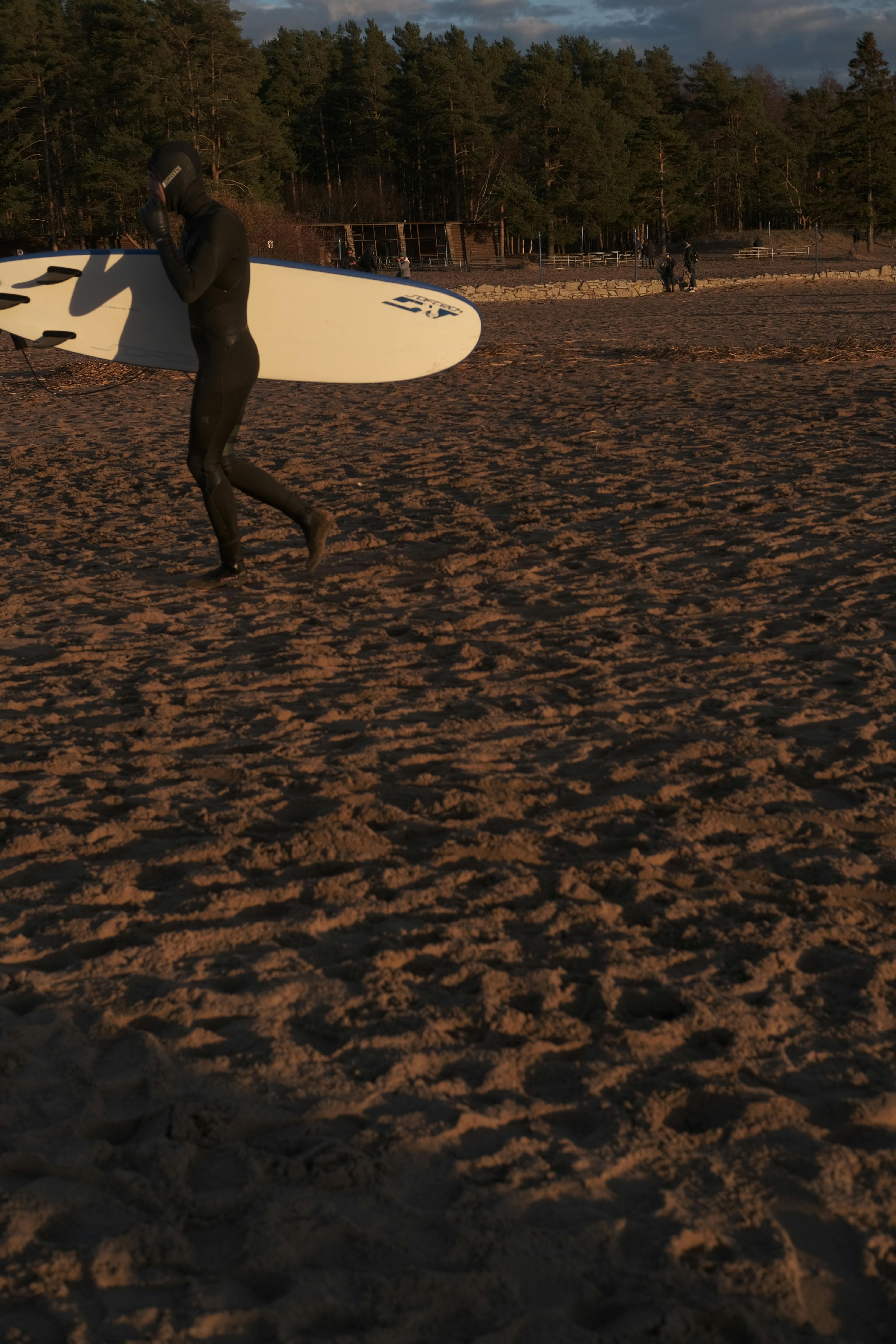 Full body side view of anonymous person with surfboard wearing swimwear strolling on sandy shore in summer evening with trees on background