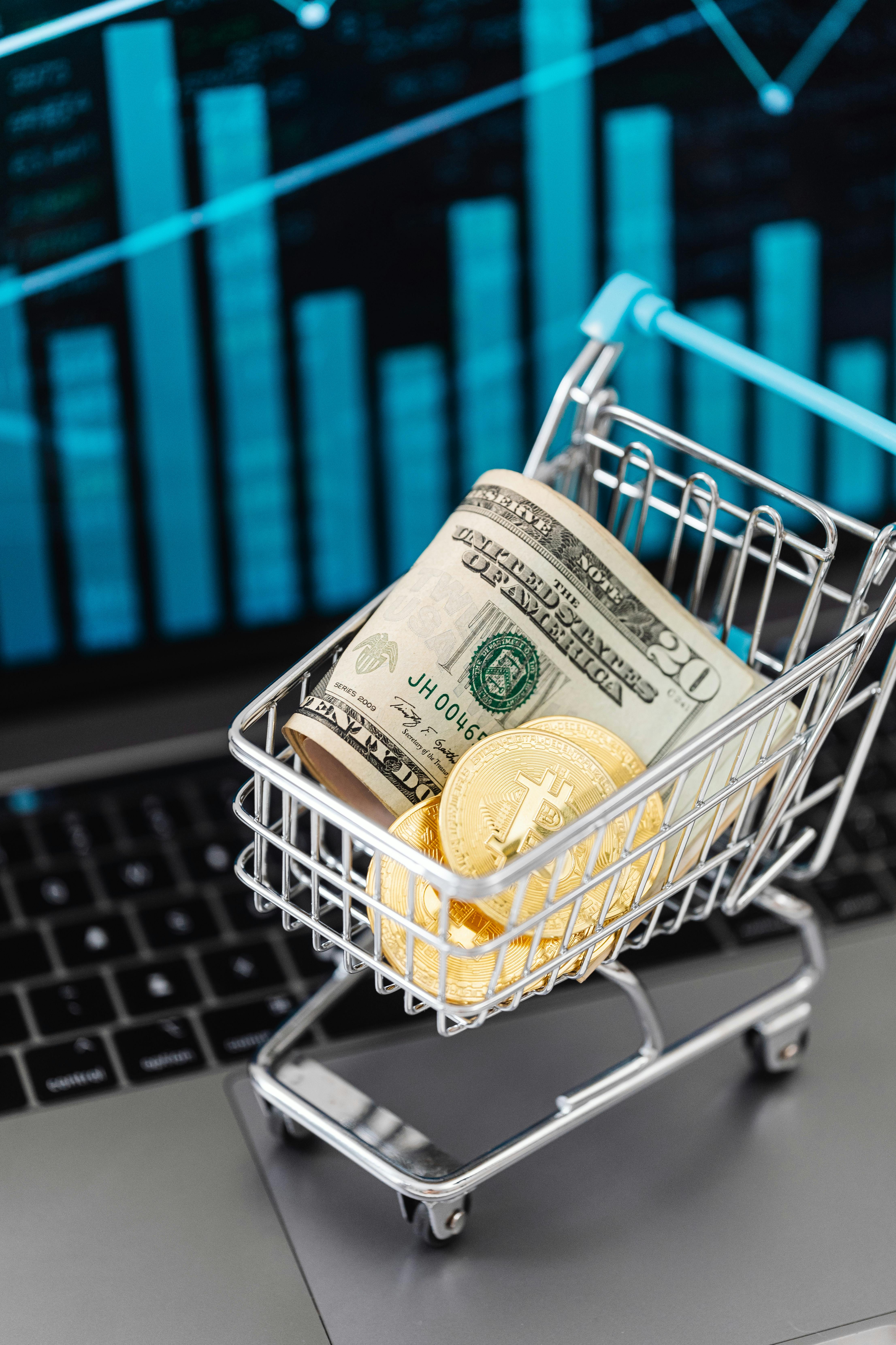 mini shopping cart with cash and gold bitcoin coins inside standing on a laptop