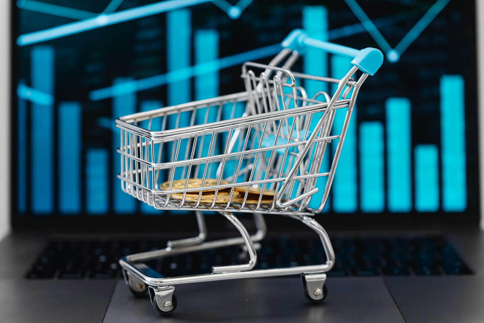 A miniature shopping cart with coins sits on a laptop displaying financial graphs, symbolizing e-commerce and online shopping trends.