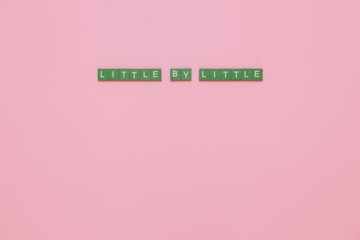 Green tiles spelling 'LITTLE BY LITTLE' on a pink background conveying motivation.