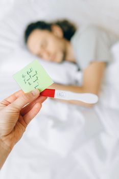 一名女子手持显示阳性的验孕棒和一张纸条，背景中有人在睡觉