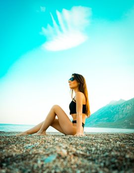 A young woman in swimwear relaxing on a sandy beach with mountains in the background.