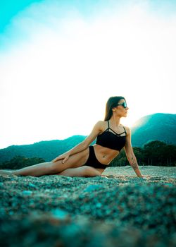 A woman in a bikini poses on a sunny beach with scenic mountains in the background.