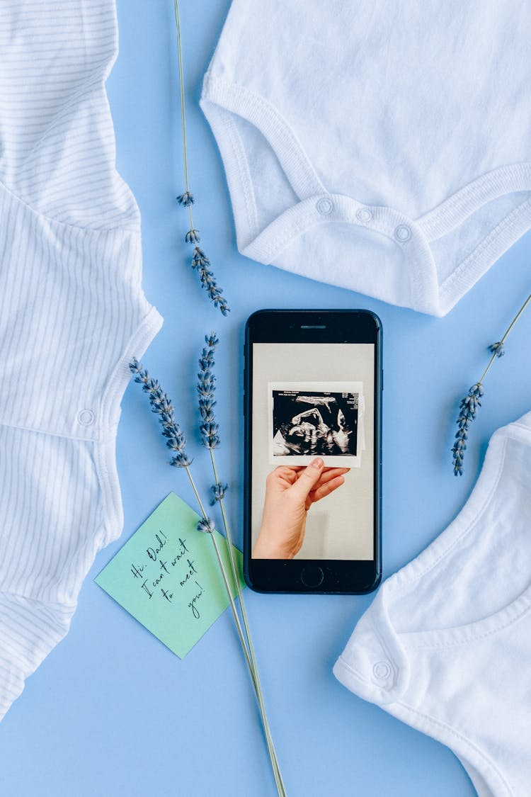 Black Cellphone With Sonogram On Screen Display Beside Baby Clothes