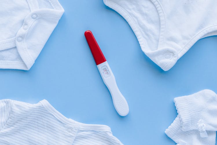 Flat Lay Photography Of Pregnancy Test And Baby Clothes On Blue Surface