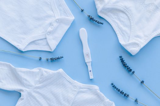 蓝色背景的妊娠测试呈阳性，婴儿衣服平放在旁