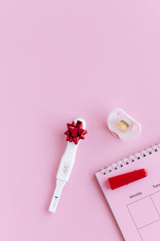 粉色背景上，平放着显示阳性结果的妊娠测试、安抚奶嘴和日历