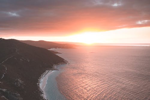 Drone view of calm ocean washing coast with mountain covered with grass against sun shining on horizon of cloudy sky at sunset time