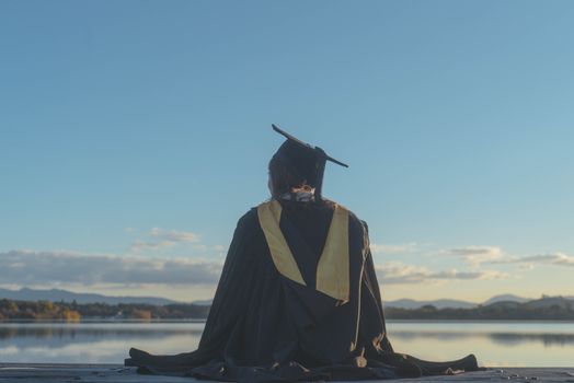 日落时分，一位戴着帽子、穿着礼服的女研究生倒映在宁静的湖面上