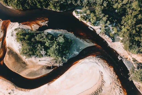 Breathtaking aerial view of boat moored on brown river flowing among sandy terrain near lush green forest in tropical countryside