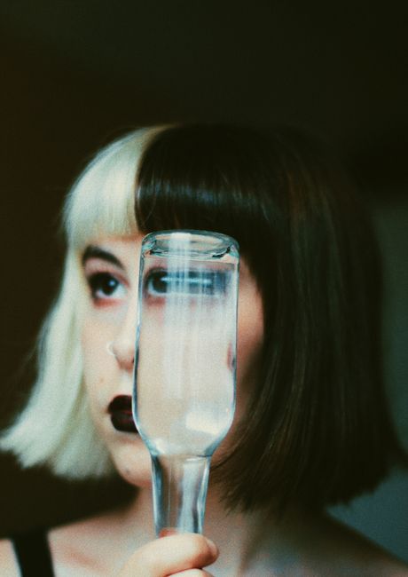 Attractive female with modern hairstyle and bright makeup cover face with glass bottle of transparent liquid and looking away