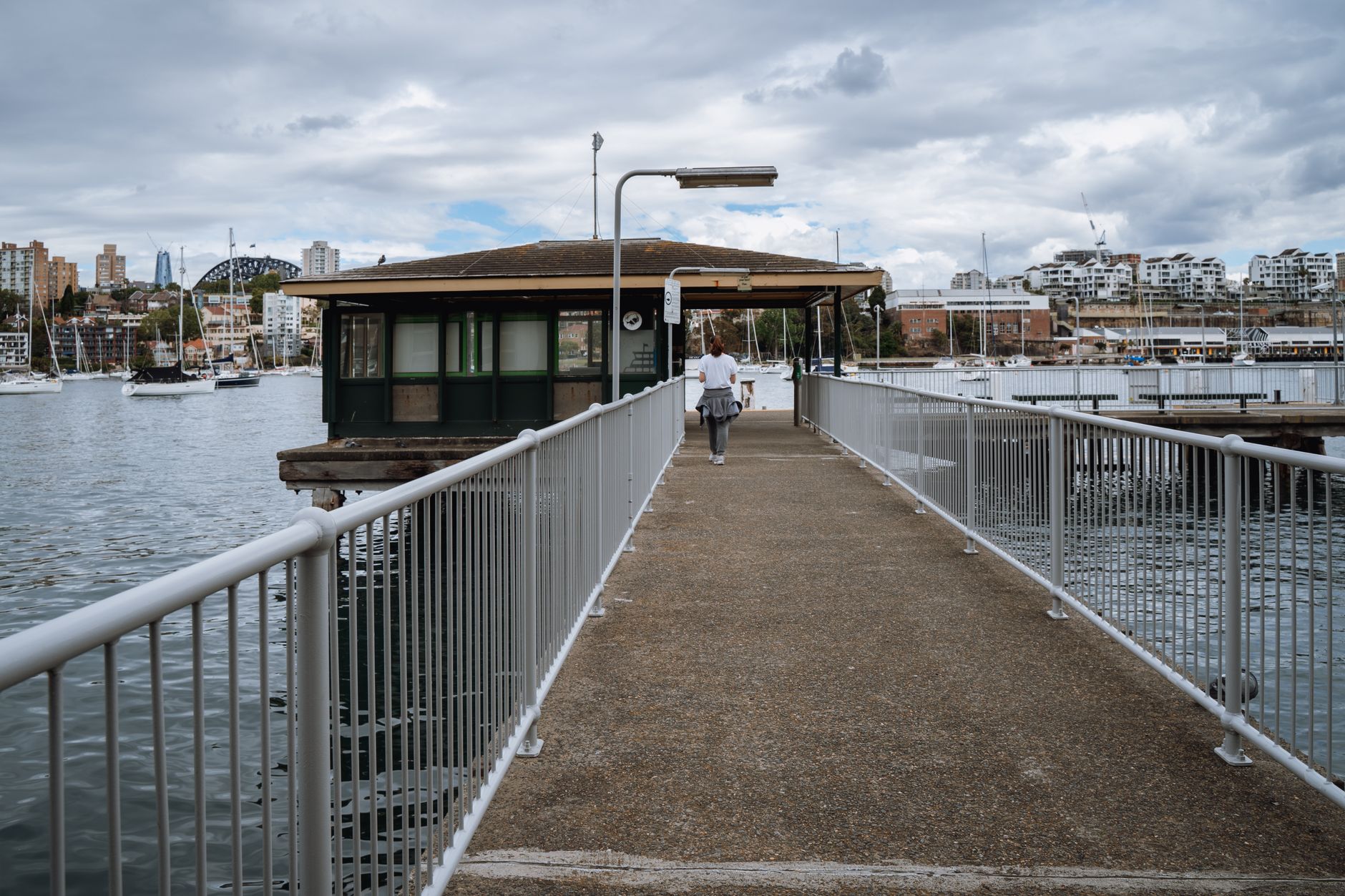 A person walks along Kurraba Point pier with the Sydney skyline ahead, close enough to belong and far enough to judge