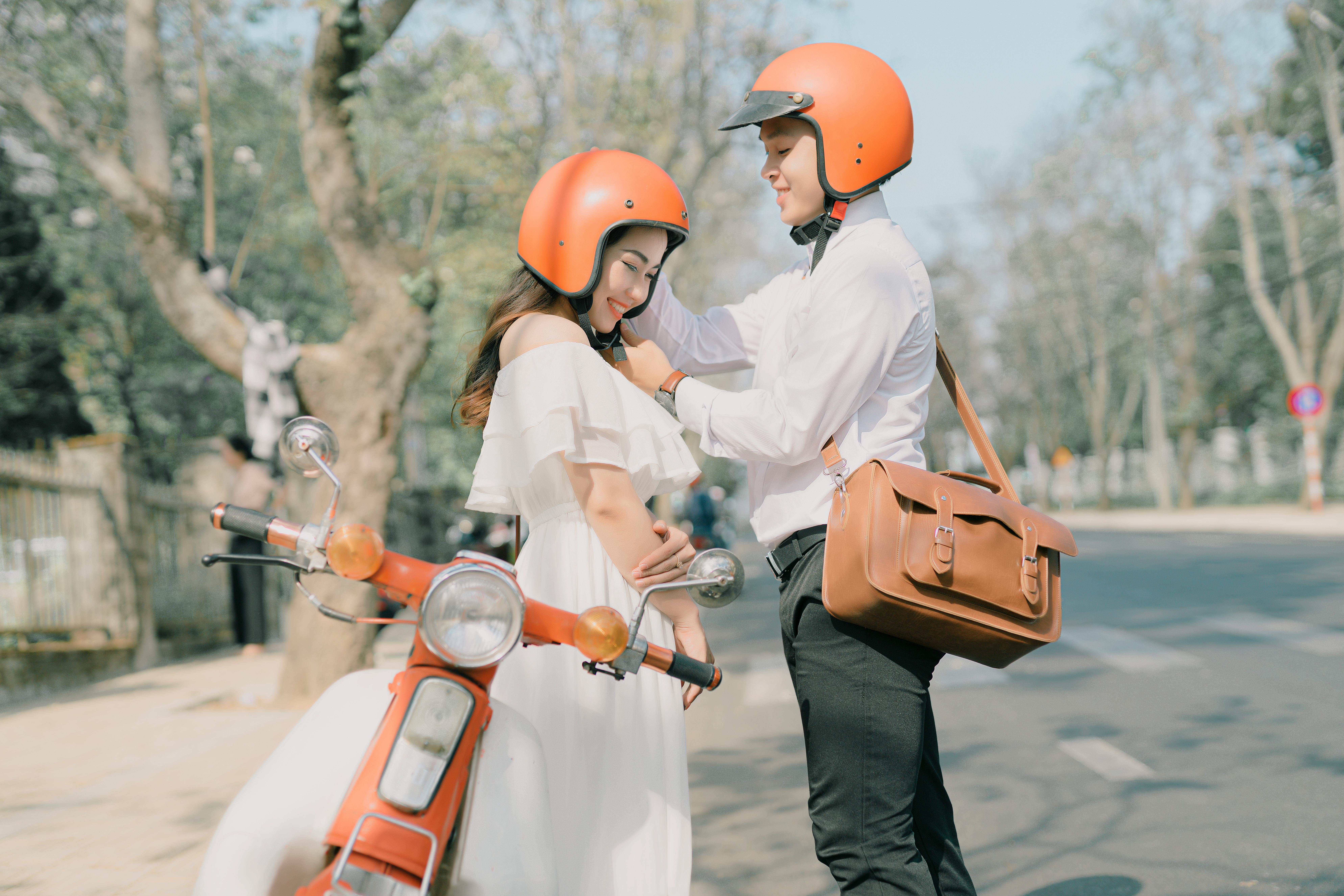 A couple embracing and adjusting helmets beside a vintage scooter on a sunny street.