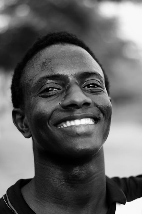 Black and white of confident young African American guy in polo shirt smiling happily while standing on street in sunlight