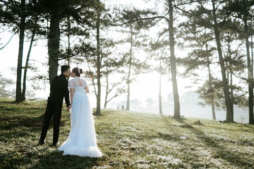 Full body of groom and bride in wedding dress holding hands and kissing each other while standing on grassy meadow in forest
