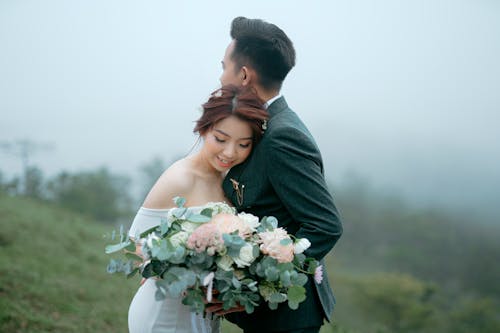 Romantic Asian couple with bouquet of flowers wearing wedding outfits embracing on grassy hill against foggy forest during holiday celebration