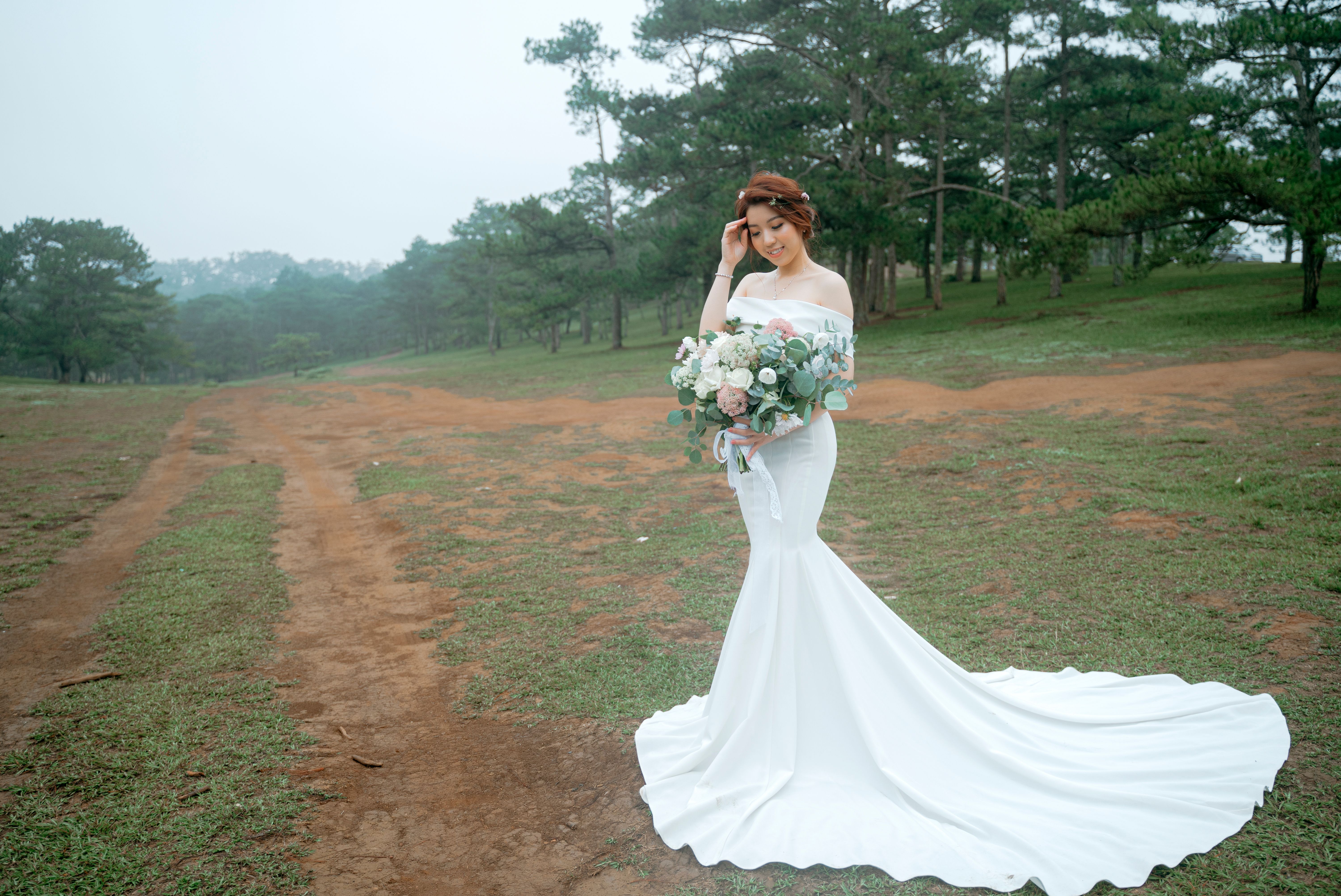 Full body of Asian woman in wedding dress touching hair while carrying bouquet of flowers in hands