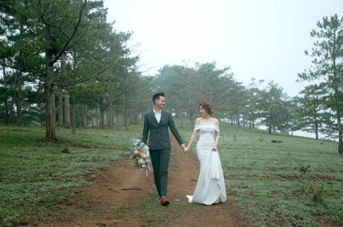 Full body of elegant newlywed couple in wedding clothes with bridal bouquet holding hands while strolling in park and looking at each other