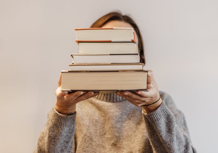 Person In Gray Sweater Carrying A Stack Of Hardbound Books