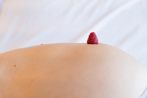 Side view of crop anonymous expectant female with bright tasty ripe raspberry on navel on white background