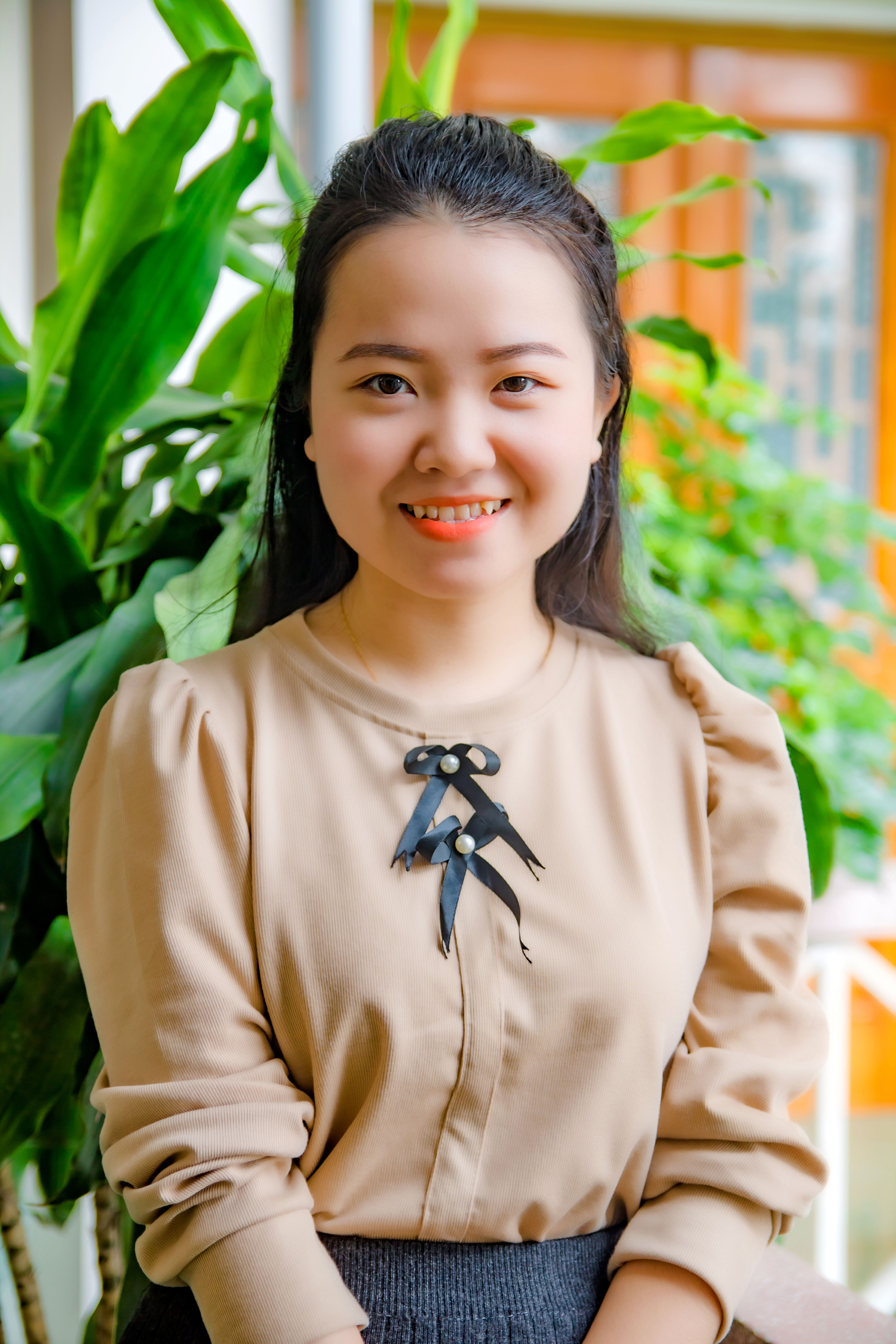 A smiling woman wearing a beige top, sitting in front of green plants. Bright and cheerful.