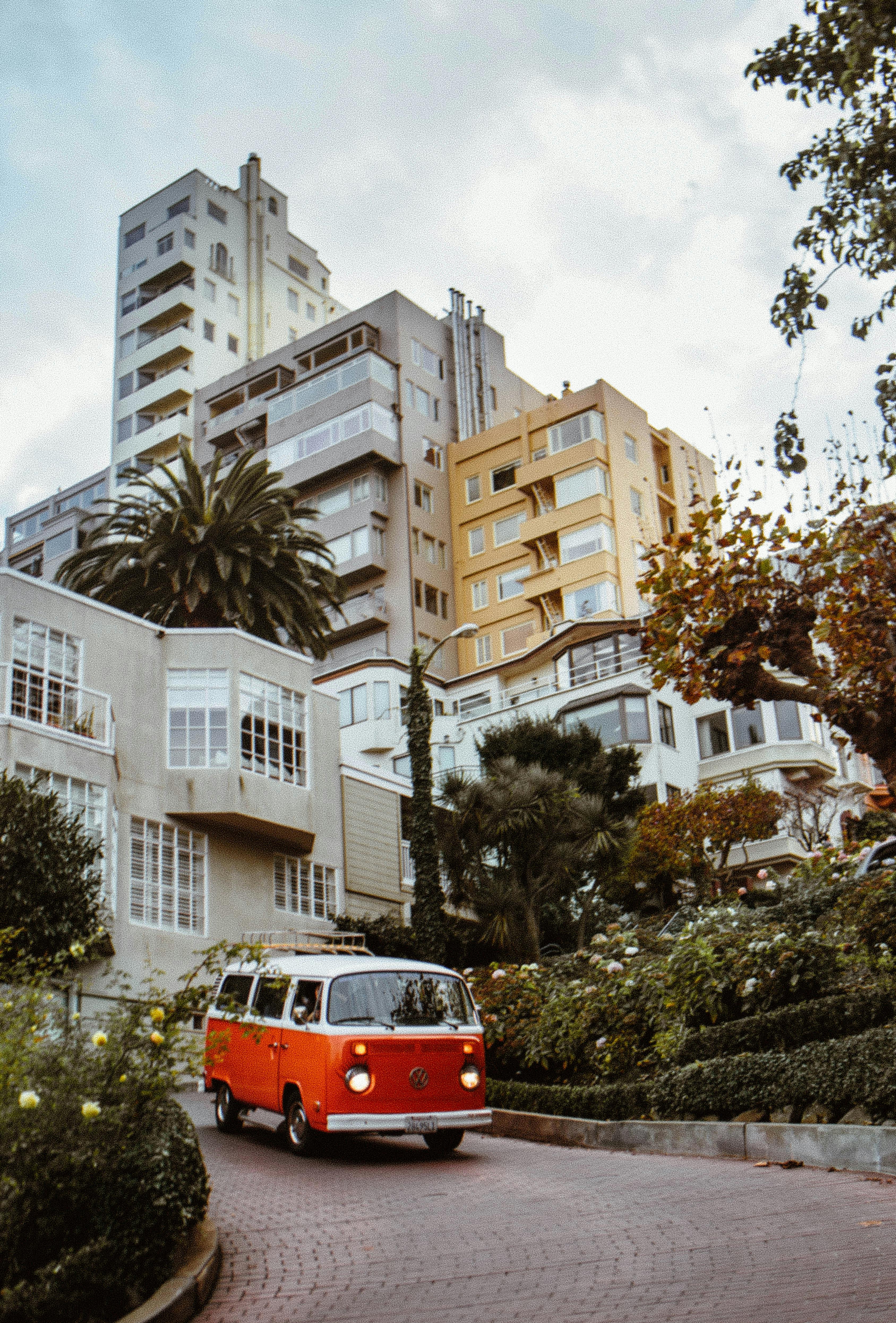 A vintage orange van drives down the iconic Lombard Street in San Francisco, surrounded by urban architecture.