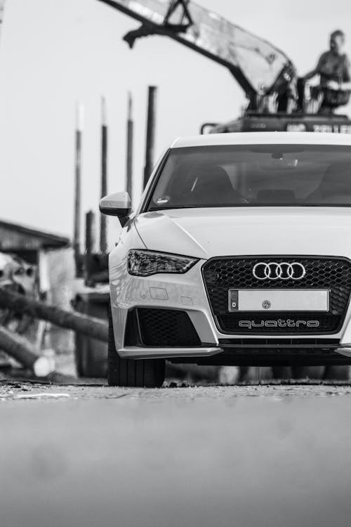 Ground level black and white of contemporary automobile riding on paved road in industrial district with construction crane and pipes on blurred background