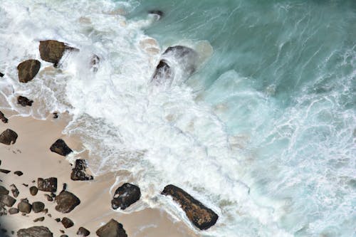 Top view of foamy waves of azure ocean washing sandy shore with boulders on island during summer day on seaside