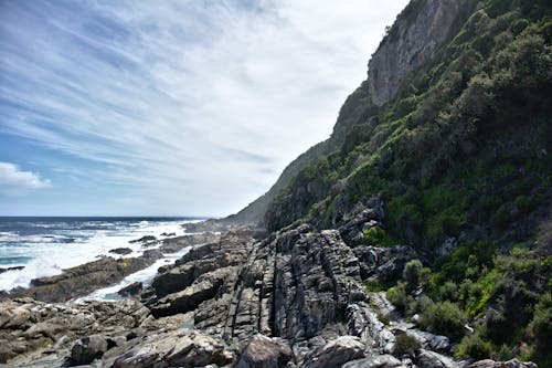 Mountain slope covered with grass located on stony shore near waving sea against cloudless sky on island on summer day