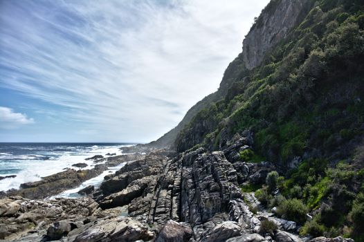 Mountain slope covered with grass located on stony shore near waving sea against cloudless sky on island on summer day