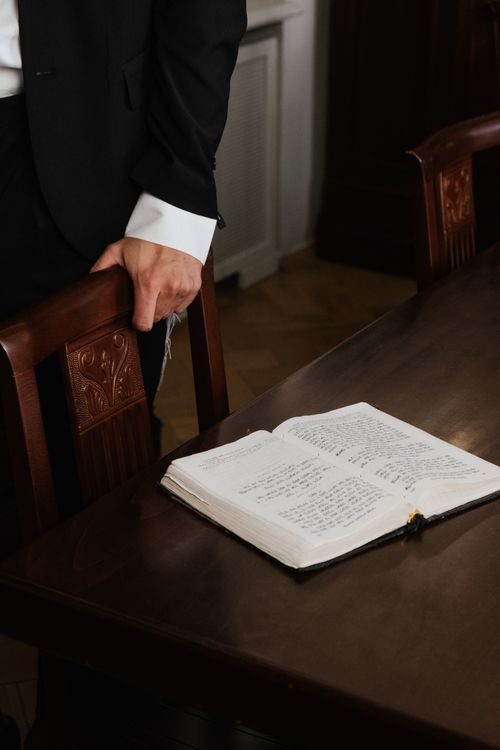 Free A businessman in a suit stands by a wooden table with an open book, implying authority. Stock Photo