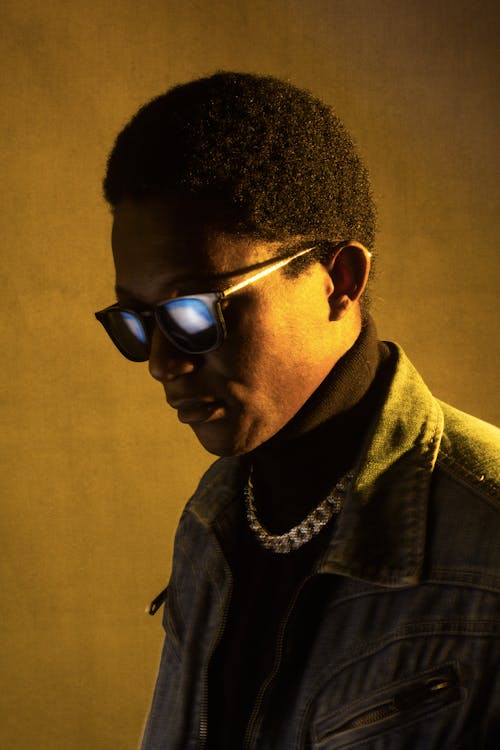 Trendy African American male with short hair wearing denim jacket and chain standing near wall with sunglasses near wall at low light