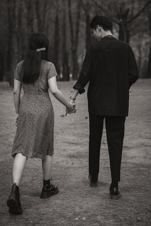 Black and white back view of unrecognizable loving couple holding hands while strolling in garden with tall trees in nature