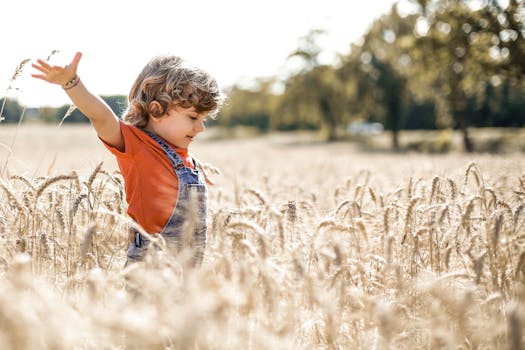 Side view of positive kid with outstretched arms in agricultural meadow with dried plants and green trees on blurred background
