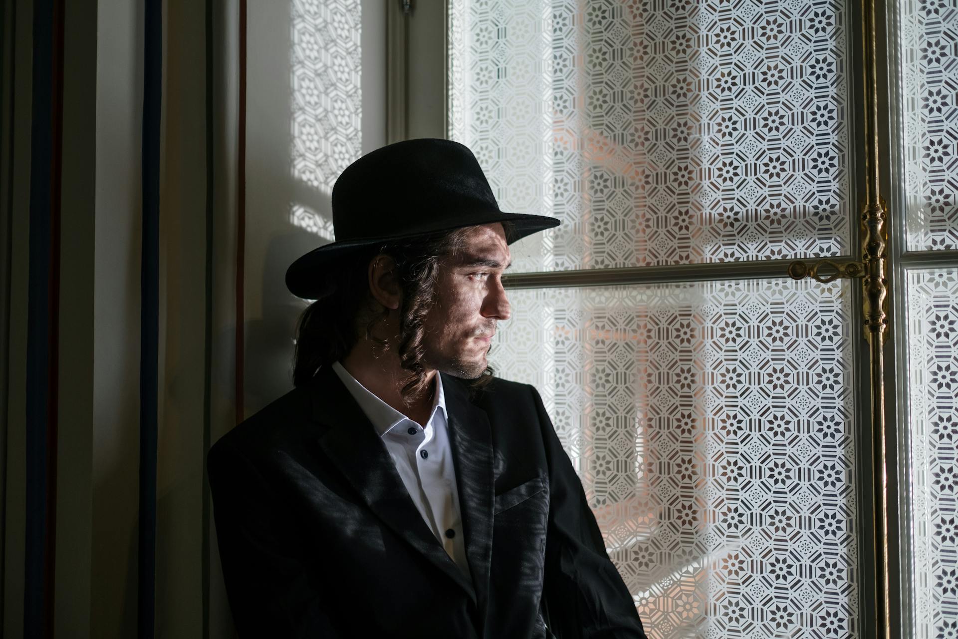 A thoughtful man in traditional Jewish clothing by a decorative window with sidelocks and a hat.