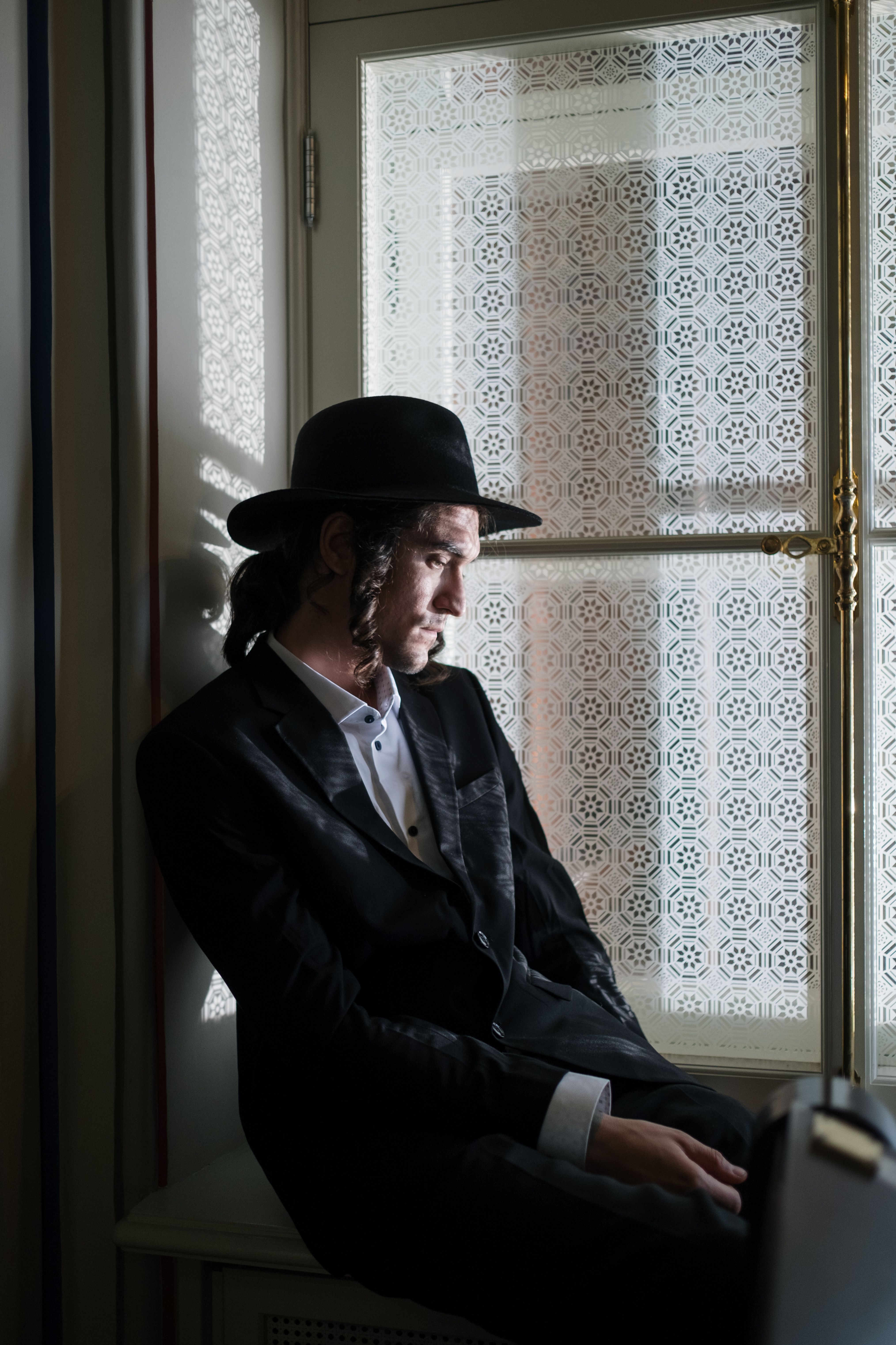 A man sits by a window in traditional Jewish attire, deep in thought.